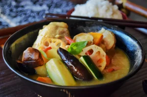 A colorful bowl of Thai green curry with chicken, lotus root, eggplant, and vegetables, garnished with basil. A bowl of rice is in the background.