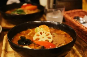 A steaming bowl of curry soup with assorted vegetables like lotus root, broccoli, and red pepper, garnished with sesame seeds, conveys warmth and flavor.