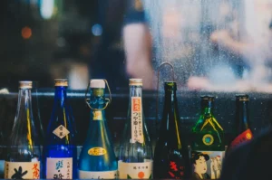 A row of assorted sake bottles with colorful labels sits on a dimly lit bar shelf, reflecting a cozy and authentic Japanese ambiance.