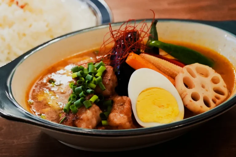 A bowl of vibrant curry soup with chunky vegetables, lotus root, a halved boiled egg, and diced green onions, evoking warmth and richness. Rice sits aside.