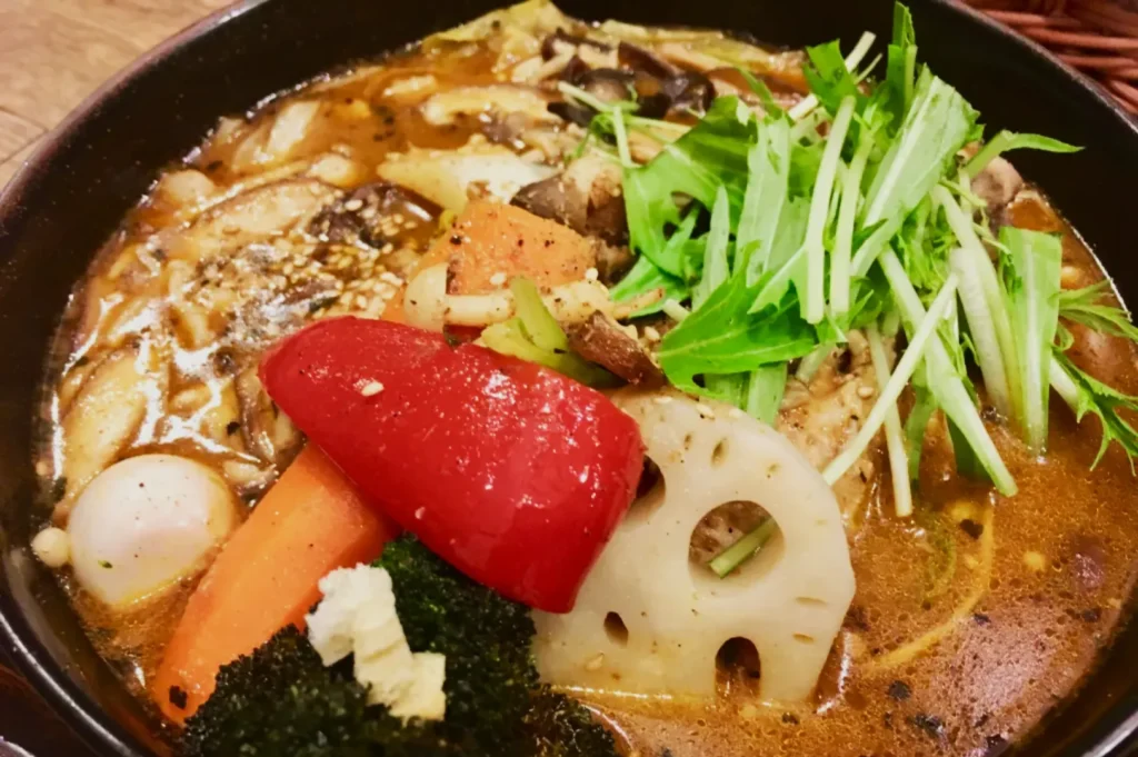 A hearty bowl of soup featuring vibrant vegetables like red peppers, lotus root, and leafy greens in a rich broth, creating a warm, comforting dish.