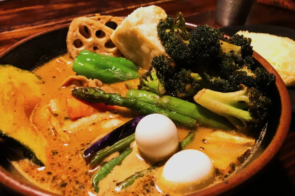A vibrant curry dish in a bowl, featuring broccoli, asparagus, peppers, tofu, lotus root, quail eggs, and a creamy sauce, creates a warm and inviting tone.