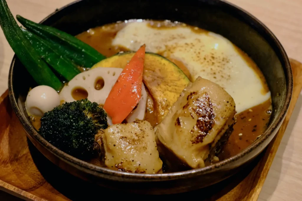 A hearty soup curry in a black bowl with grilled vegetables like carrot, lotus root, broccoli, and okra, topped with molten cheese, exuding warmth.