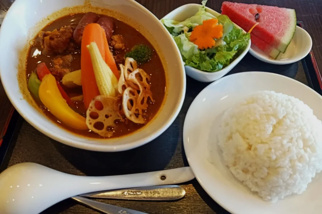 A bowl of colorful curry soup with vegetables like carrot, bell peppers, and lotus root; served with white rice, a fresh salad, and watermelon slices.