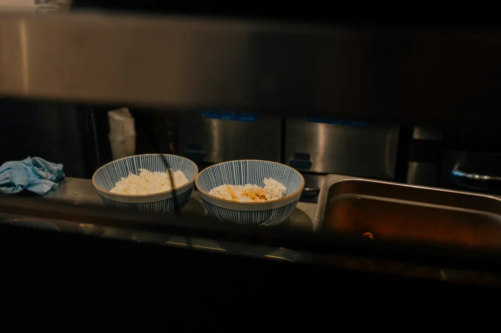Two blue-striped bowls filled with rice and other ingredients sit on a kitchen counter. A blue cloth is nearby, with a dim, industrial background.