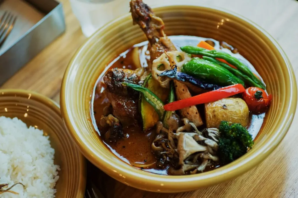 A warm, vibrant bowl of curry soup with chicken, assorted vegetables, and colorful peppers sits next to a plate of white rice on a wooden table.
