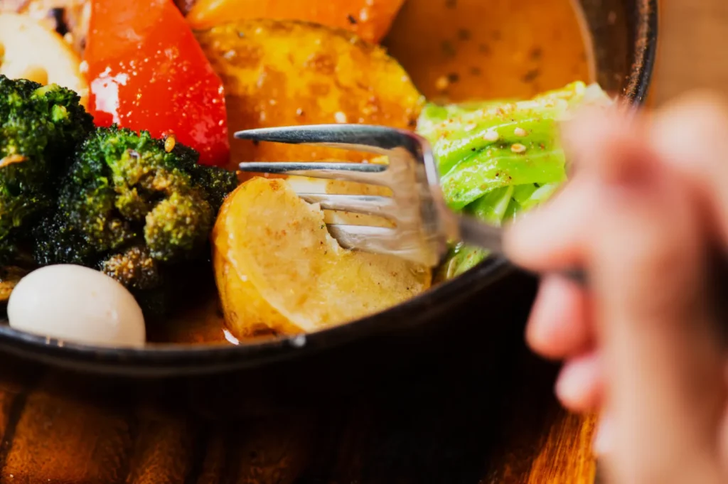 Close-up of a bowl with vibrant vegetables, including broccoli, red bell pepper, and cabbage. A fork is piercing a potato slice; warm and inviting.