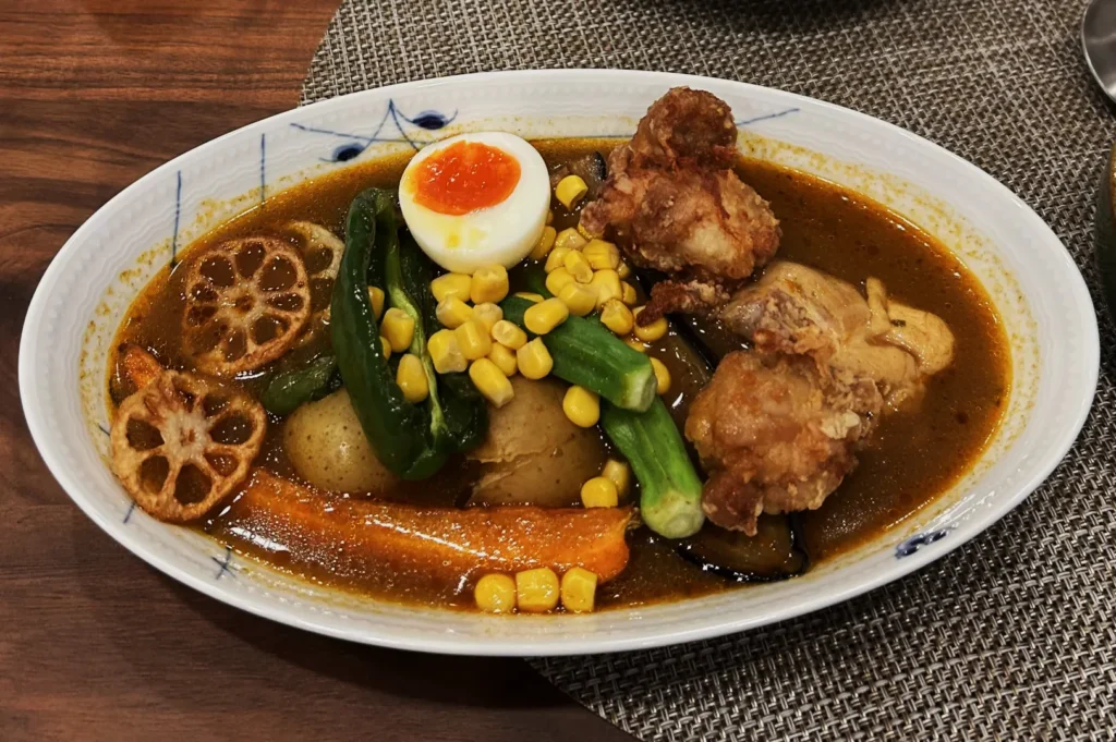 A bowl of Japanese curry with crispy fried chicken, a boiled egg, lotus root, vegetables, and corn on a wooden table, conveying warmth and comfort.