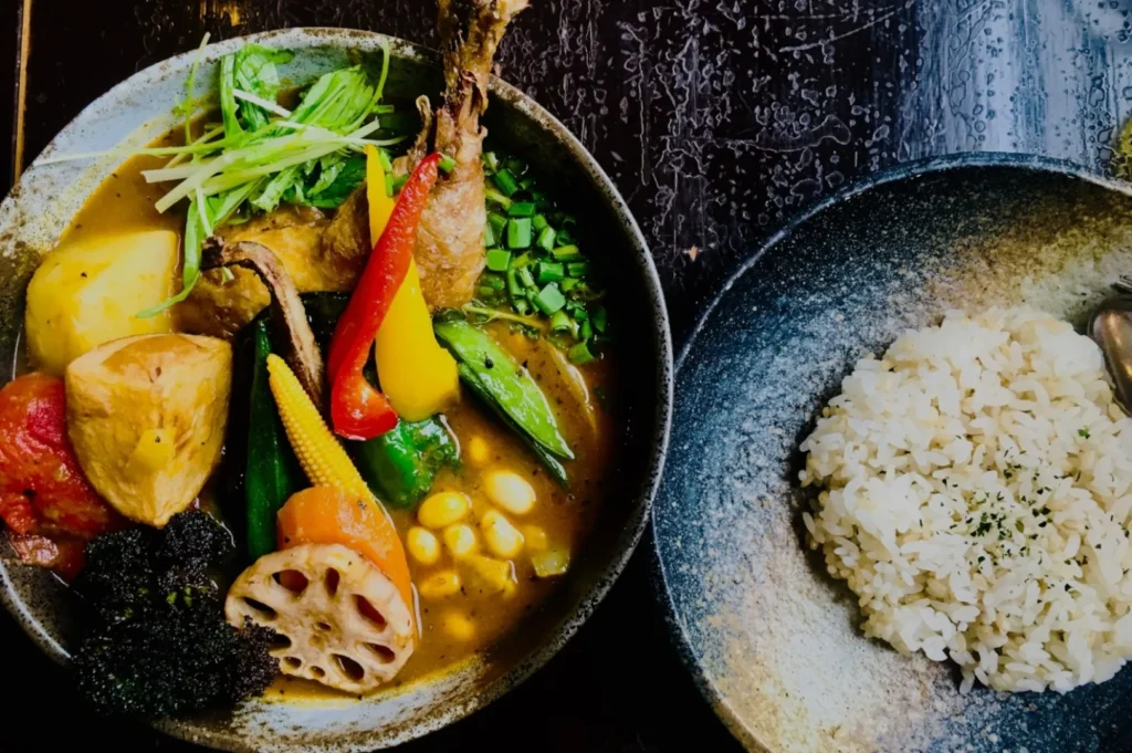 A colorful bowl of Japanese curry with vibrant vegetables and a fried drumstick, next to a plate of seasoned rice. The dish looks appetizing and aromatic.