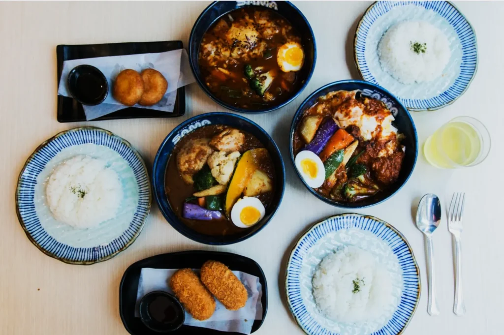 A selection of Japanese curry dishes with rice, colorful vegetables, and boiled eggs in blue bowls, alongside fried snacks and a glass of juice.