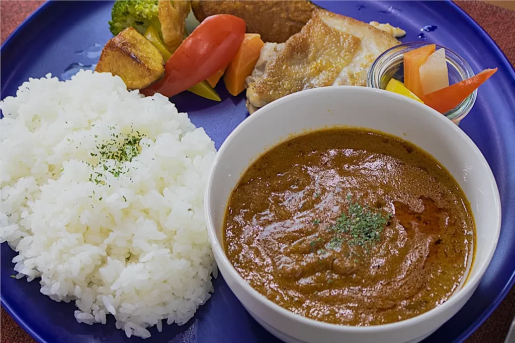 A blue plate with white rice garnished with herbs, a bowl of brown curry, grilled chicken, and colorful vegetables, conveying a hearty meal.