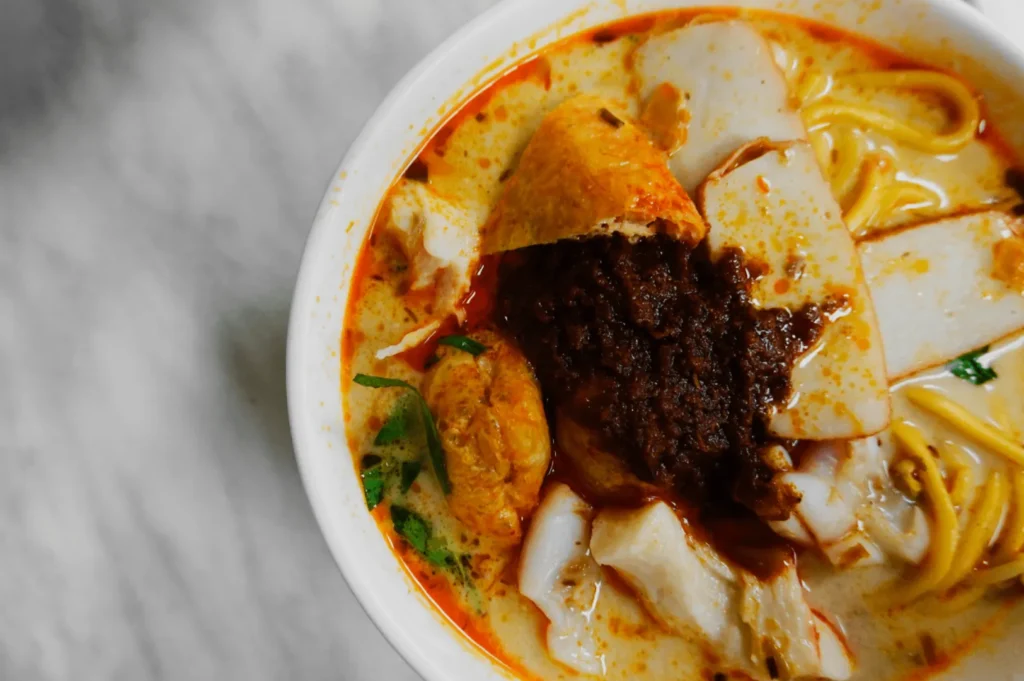 A bowl of creamy laksa with noodles, topped with fish cake slices, tofu, fresh herbs, and a dollop of dark, spicy sambal paste on a marble surface.