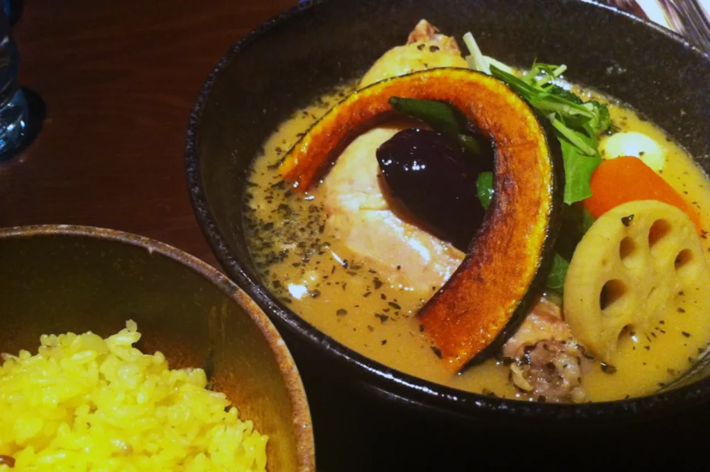 A bowl of creamy soup curry filled with chicken, pumpkin slice, eggplant, and lotus root sits beside a bowl of vibrant yellow rice on a wooden table.