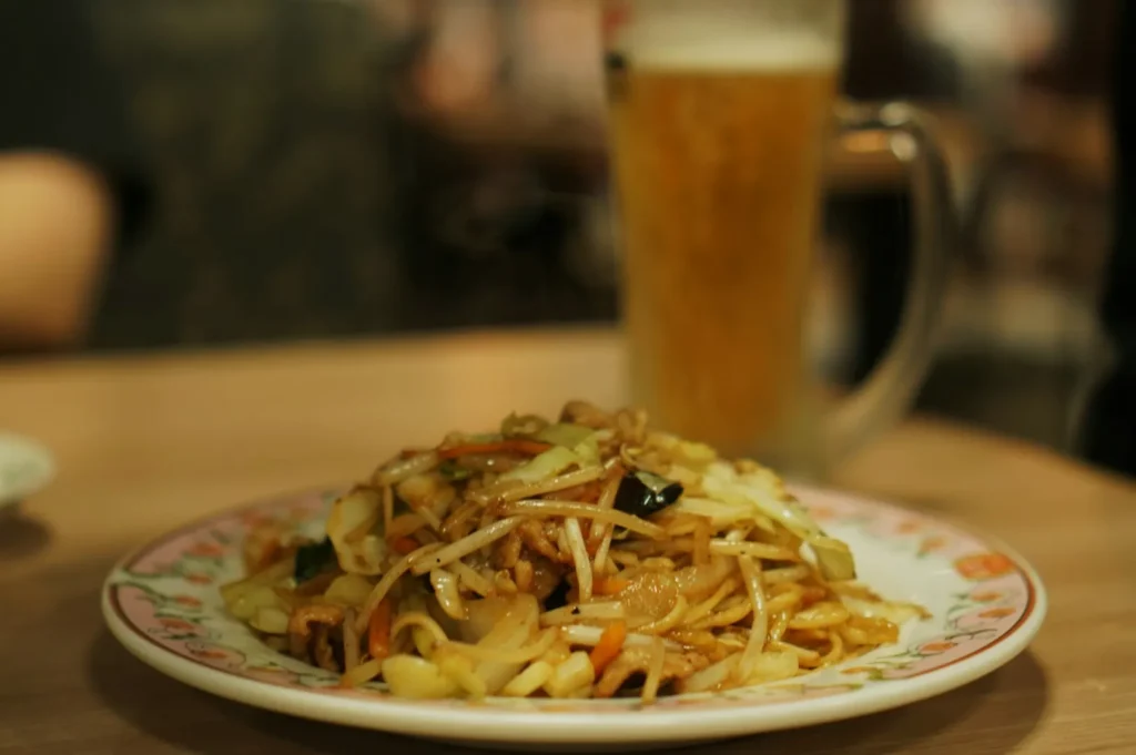 Plate of stir-fried noodles with vegetables and meat on a table, paired with a glass of frothy beer in the background, creating a cozy dining ambiance.