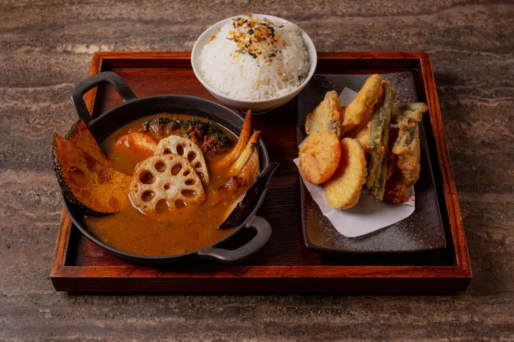 A tray with vibrant vegetable curry, white rice topped with herbs in a bowl, and assorted golden tempura on a plate, set on a rustic table.