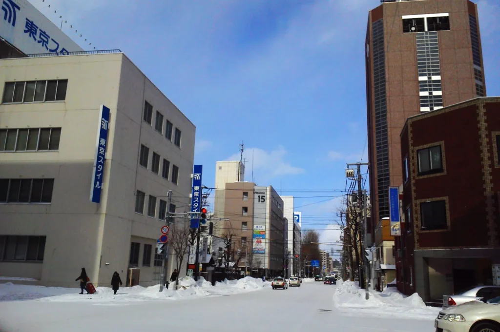 Snow-covered urban street with several buildings lining both sides under a bright blue sky. People walk along icy sidewalks, creating a cold, bustling city scene.