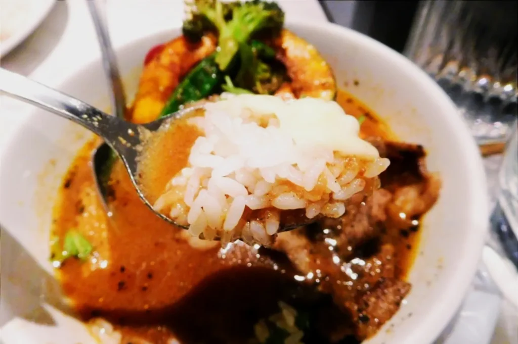 A spoonful of rice and curry hovers over a bowl filled with rich brown curry, broccoli, and grilled vegetables, exuding warmth and comfort.