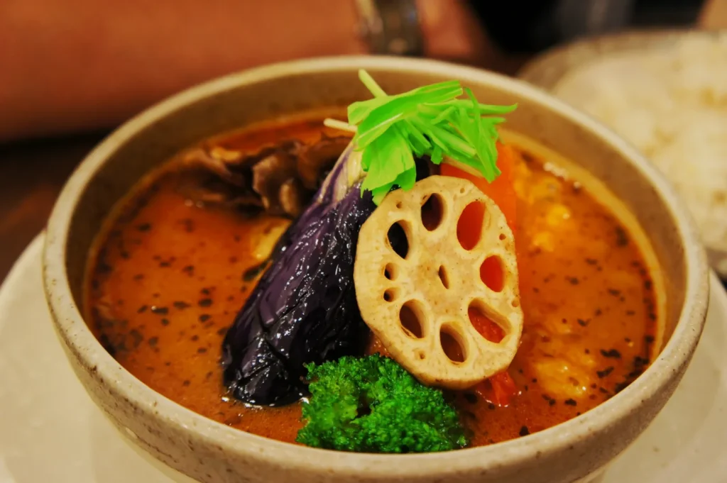 A vibrant bowl of vegetable curry soup featuring a lotus root slice, purple eggplant, broccoli, and fresh greens, creating a warm, appetizing feel.