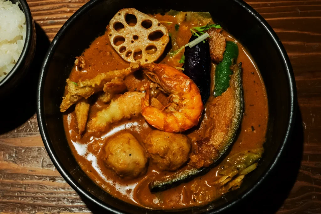 Bowl of Japanese curry soup with shrimp, lotus root, vegetables, and rice on the side. Warm, hearty, and savory with vibrant colors.