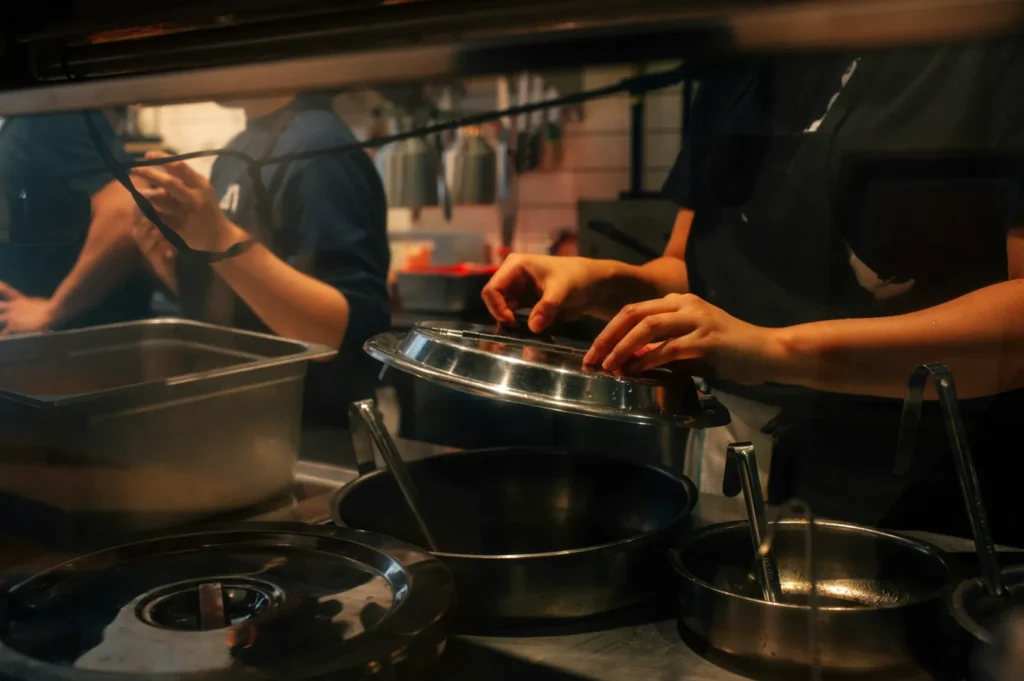 Hands lift a metal lid off a pot in a bustling kitchen. Utensils and cookware are visible, with a focused, busy atmosphere.