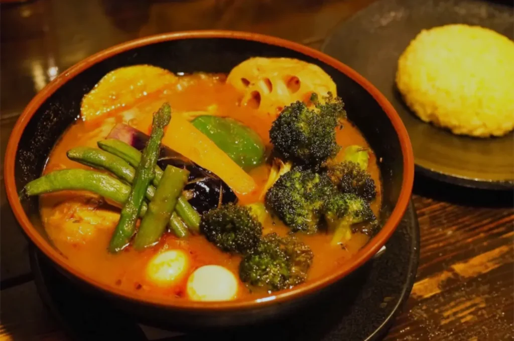 A bowl of colorful vegetable curry with broccoli, carrots, lotus root, and beans in rich sauce. A serving of rice is on a black plate nearby.