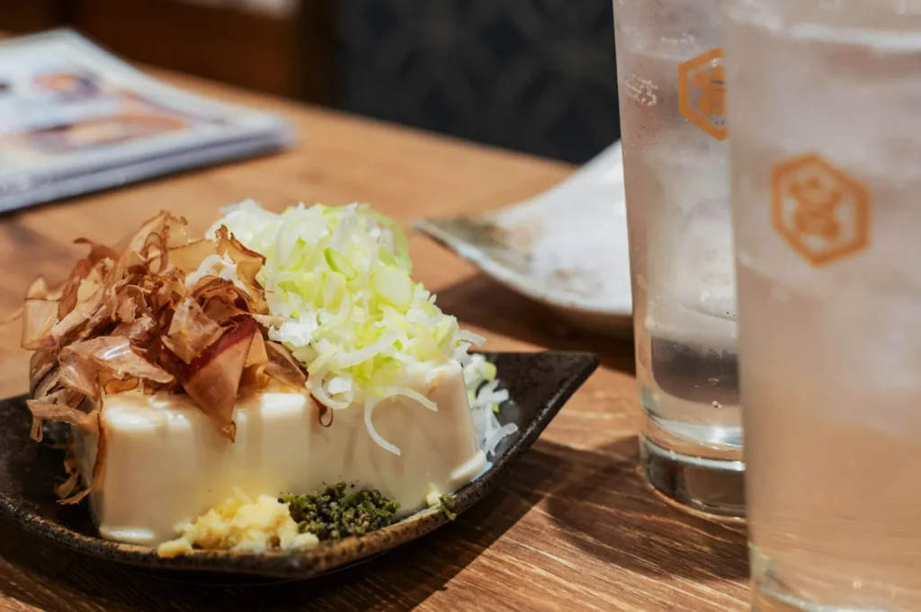 A square piece of tofu topped with bonito flakes, chopped green onions, and ginger sits on a dark plate. Two tall glasses are nearby on a wooden table.