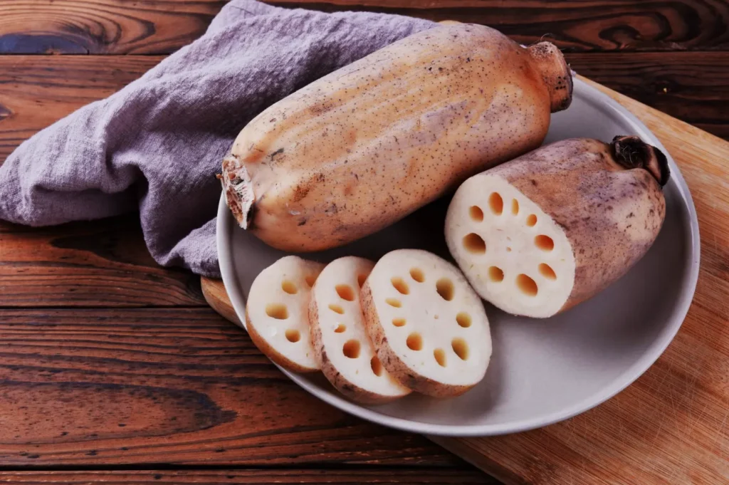Lotus root on a plate with wooden background, showcasing whole and sliced pieces. A lavender cloth adds a rustic kitchen feel.