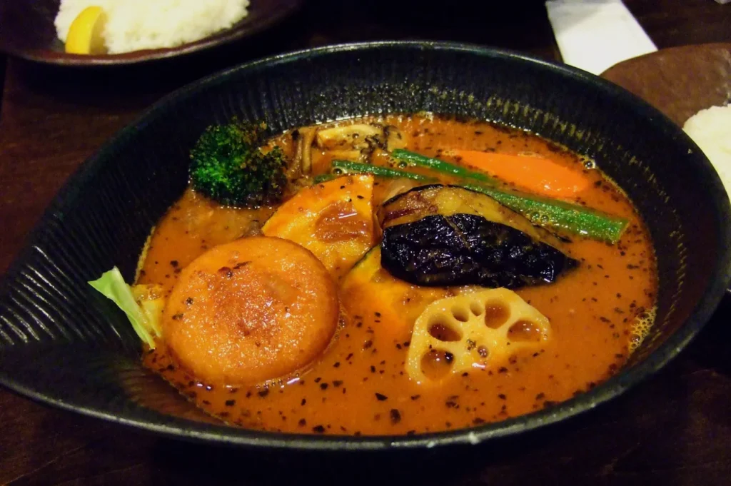 A black bowl of vibrant curry soup topped with assorted vegetables like broccoli, carrots, and lotus root, served beside a plate of white rice.