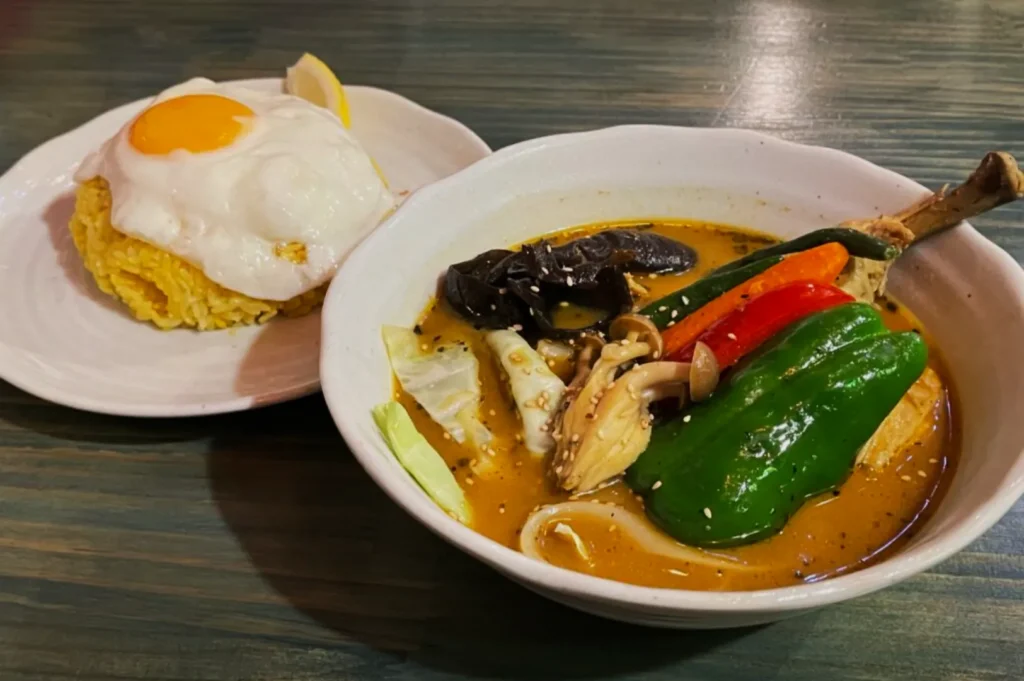 A white bowl of curry soup with a chicken drumstick, bell peppers, mushrooms, and cabbage sits beside a plate of saffron rice topped with a sunny-side-up egg.