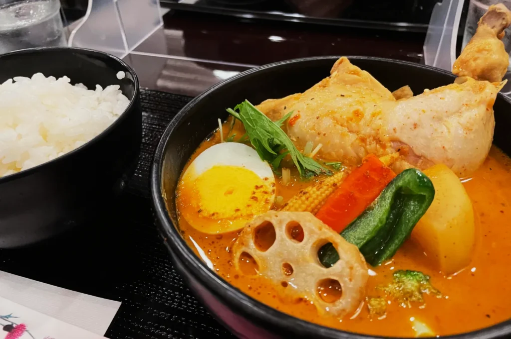 A bowl of vibrant orange curry soup with chicken, boiled egg, vegetables, and lotus root, next to a bowl of white rice on a dark table.