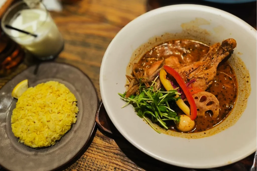 A bowl of spicy curry with chicken drumstick, lotus root, and greens, paired with a plate of yellow rice and a lemon wedge. A drink in the background.