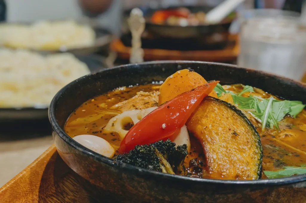 A vibrant bowl of vegetable curry soup with sliced pumpkin, red pepper, broccoli, and greens. The setting is cozy, with another dish blurred in the background.