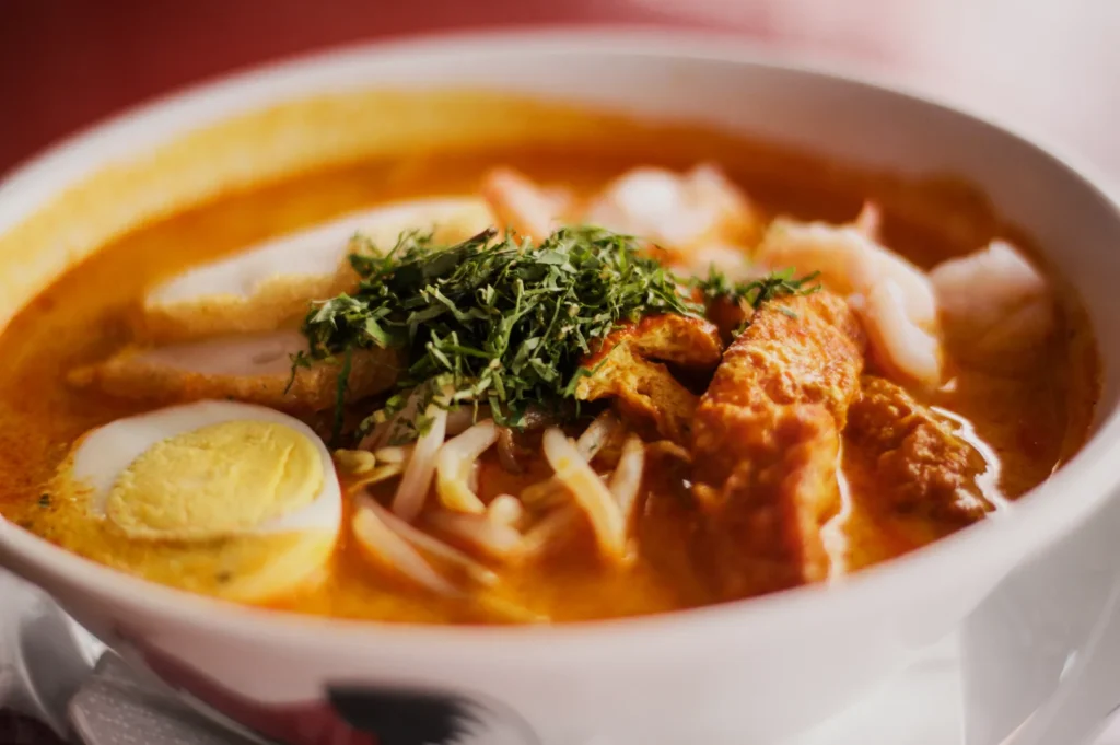 A bowl of laksa soup with a rich, orange broth, featuring half a boiled egg, shrimp, tofu, bean sprouts, and herbs, creating a hearty and inviting dish.