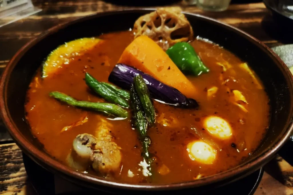 A vibrant bowl of soup with assorted vegetables, including carrots, green beans, eggplant, and lotus root, in a rich, spicy broth on a wooden table.