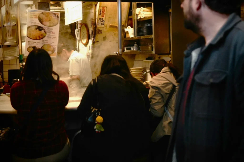 A cozy, bustling ramen shop with a chef cooking behind a steaming counter. Three customers sit closely, absorbed in the warm, aromatic atmosphere.