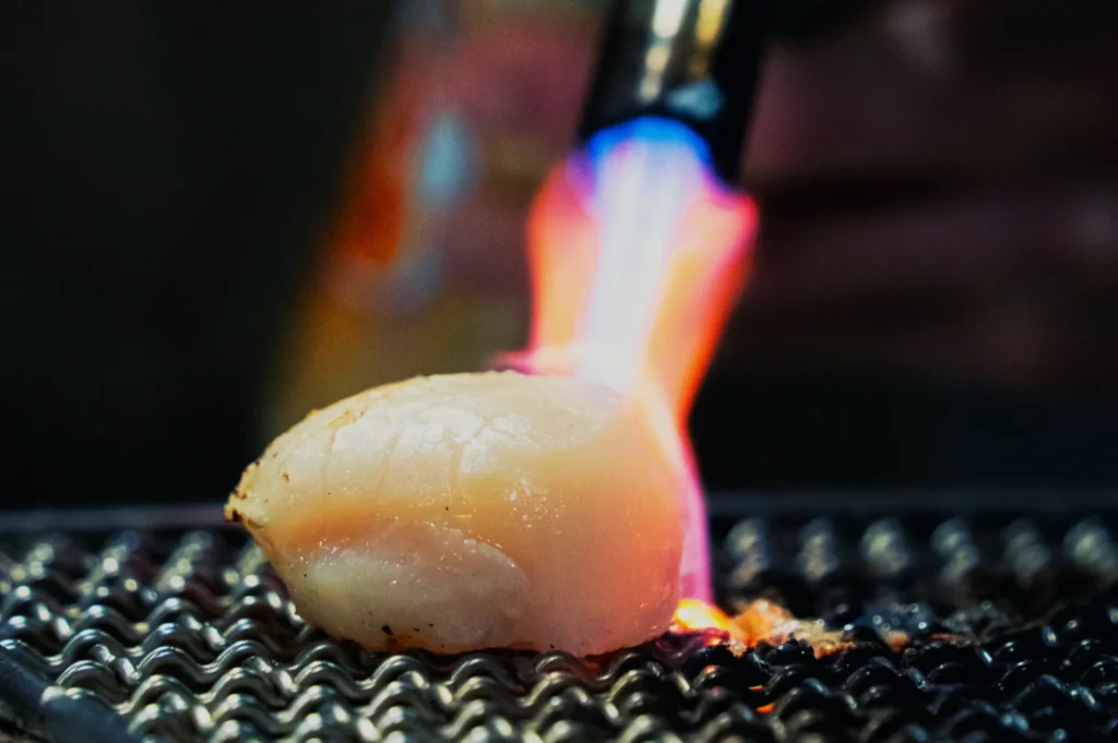 A scallop being torched on a grill with bright blue and orange flames. The scene captures the cooking process with intense heat and a focused, dynamic energy.