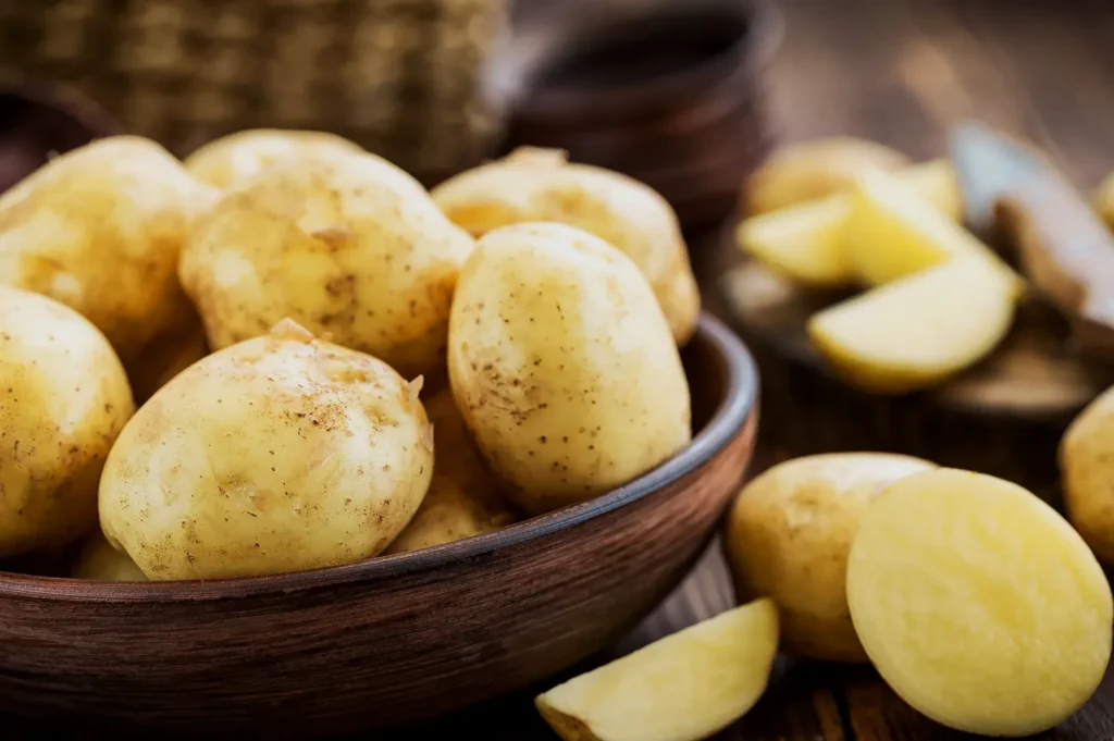 A wooden bowl filled with fresh, unpeeled potatoes on a rustic table; some potatoes are sliced, creating a homely and earthy atmosphere.