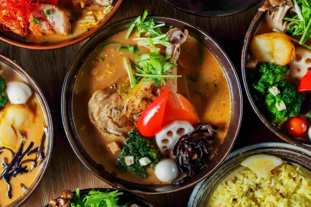 A vibrant bowl of curry soup is filled with assorted vegetables, herbs, and a chicken drumstick. Surrounding bowls hold more colorful dishes, evoking warmth and richness.