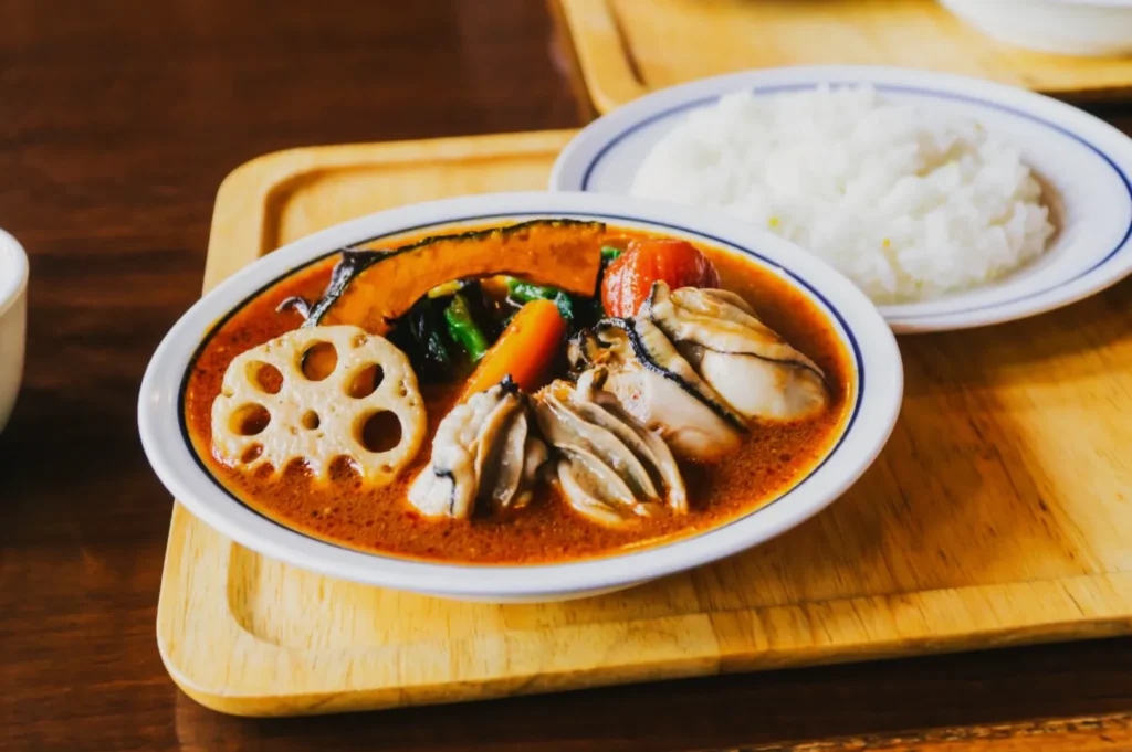 A vibrant curry dish with sliced lotus root, mussels, and vegetables in red sauce, served on a wooden tray beside a plate of white rice.
