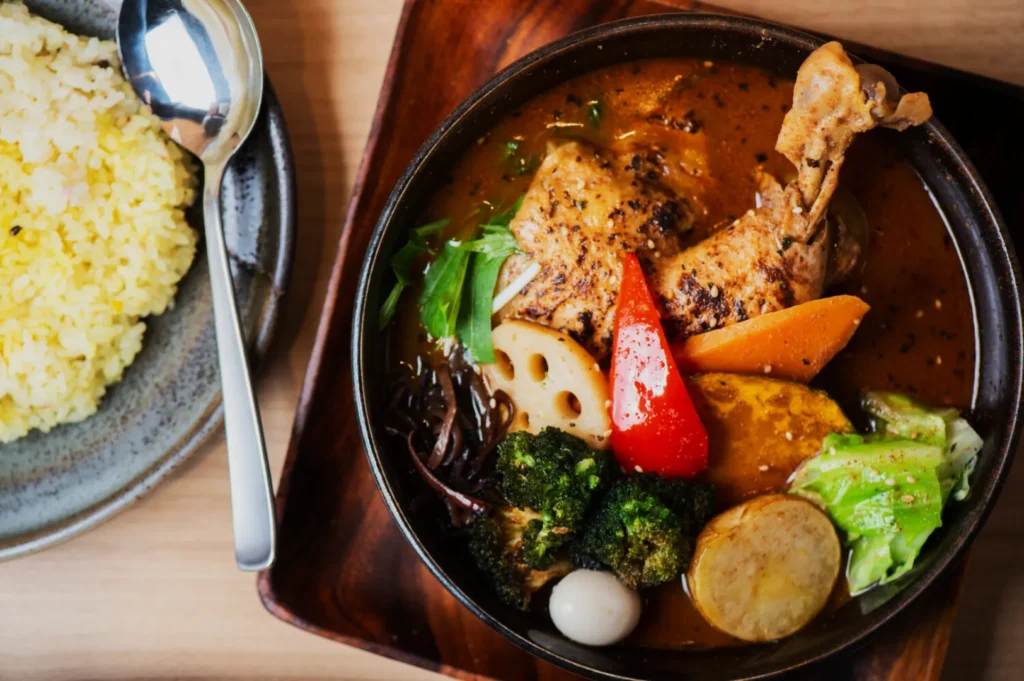 A bowl of vibrant Japanese curry with chicken leg, assorted vegetables, including broccoli and carrot, next to a plate of yellow rice with a spoon.