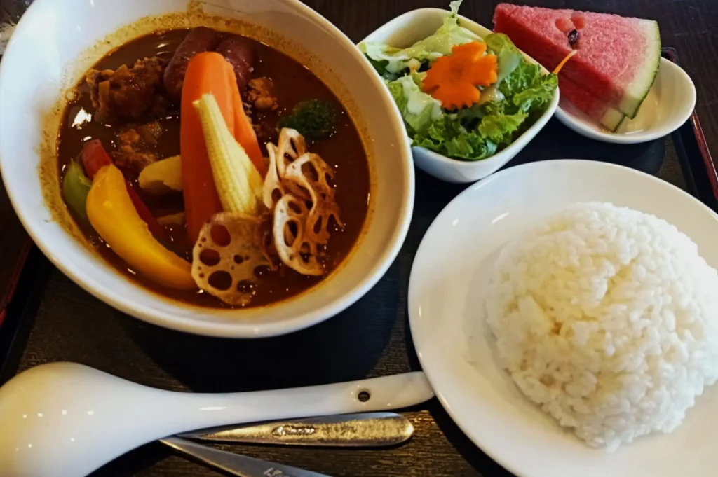 A vibrant bowl of curry with carrots, bell peppers, and lotus root sits beside a plate of white rice, a fresh salad, and watermelon slices.