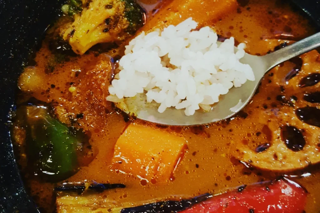 A spoon with white rice rests atop a colorful Japanese curry soup, featuring vibrant vegetables like carrots, broccoli, and lotus root in a rich broth.