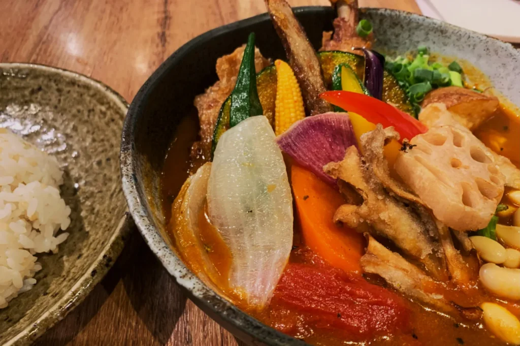 A vibrant bowl of Japanese curry soup filled with assorted colorful vegetables, including sliced lotus root, carrots, and peppers. A side of white rice sits on a rustic plate. The setting conveys a warm, inviting meal.