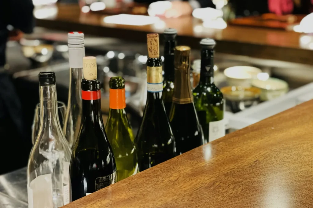 A row of various wine bottles with different colored caps and corks on a wooden bar top. The background is softly blurred, suggesting a cozy bar scene.