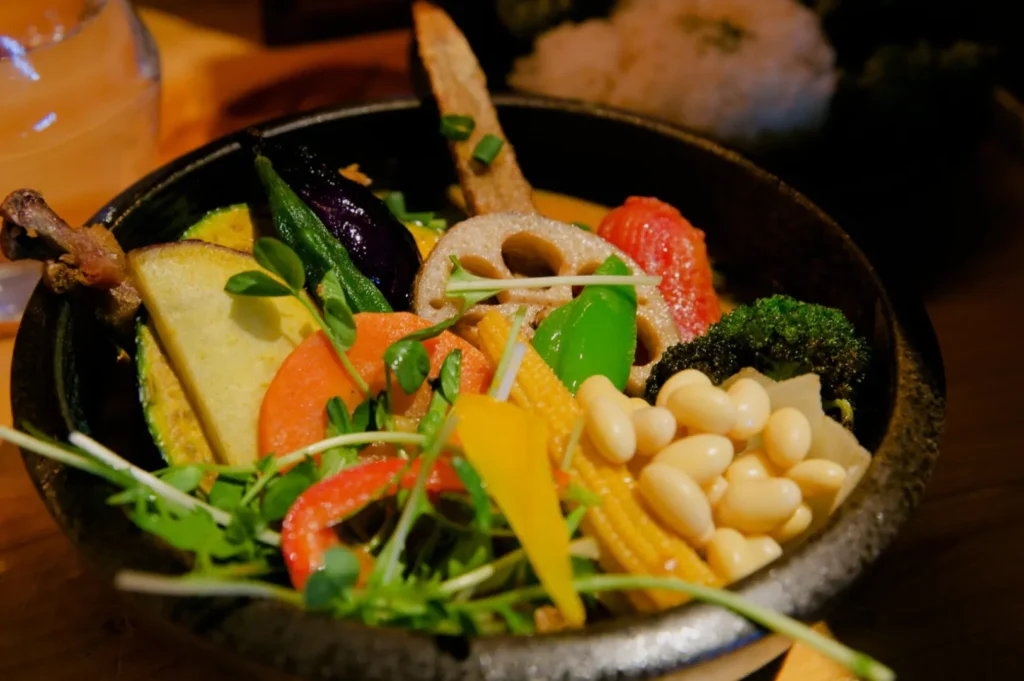 A colorful vegetable dish in a black bowl featuring lotus root, bell peppers, broccoli, beans, and microgreens, creating a fresh, vibrant appeal.