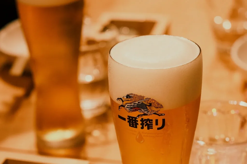A frothy glass of beer with Japanese text and a dragon logo on it. The background features blurred glasses and plates, creating a warm and inviting atmosphere.