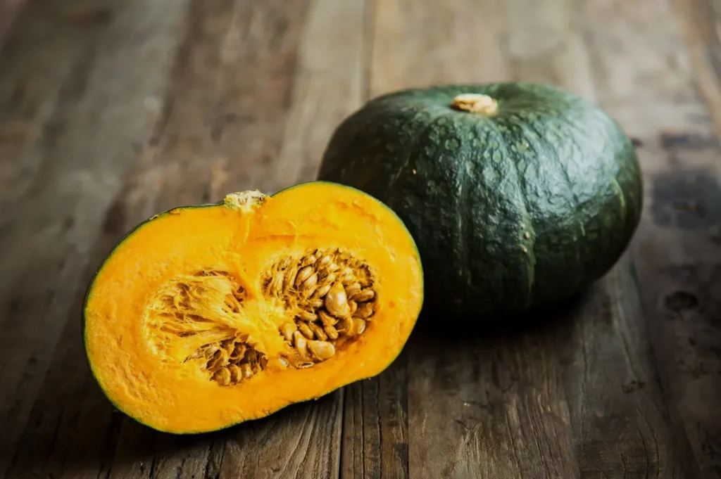 A whole dark green kabocha squash and a halved one displaying vibrant orange flesh and seeds, resting on a rustic wooden surface.
