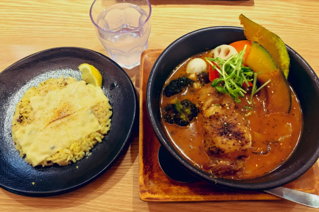 A vibrant meal features a black bowl of rich curry with chicken, vegetables, and greens, alongside a plate of cheesy yellow rice. A glass of water is nearby.