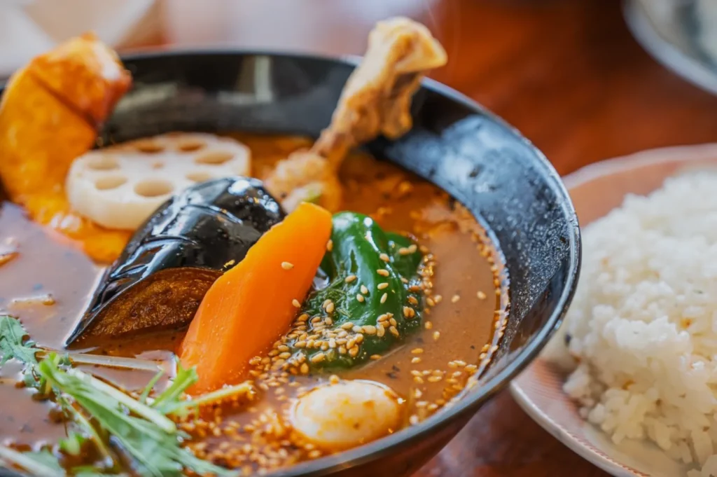 A vibrant bowl of Japanese curry soup with eggplant, carrot, pumpkin, lotus root, and a chicken leg, garnished with sesame seeds. Steaming rice is served on the side.