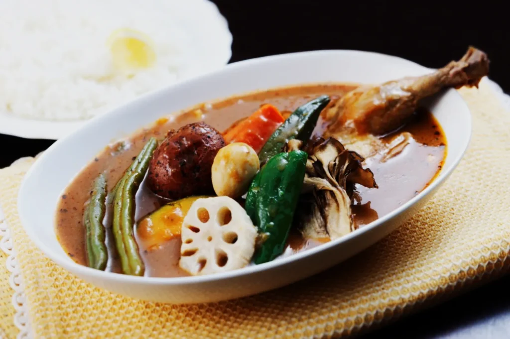 A bowl of colorful Japanese curry soup with chicken, assorted vegetables like lotus root, carrot, and mushroom on a yellow mat. Rice with a lemon is in the background.